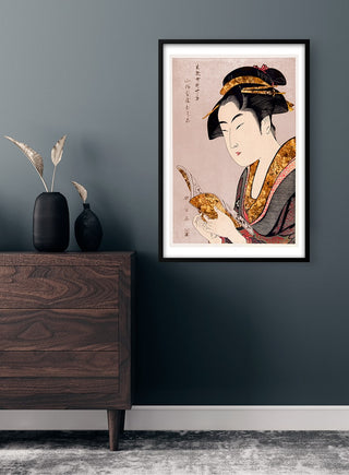 Framed poster of a traditional Japanese woman reading, displayed on a modern dark wall above wooden furniture.