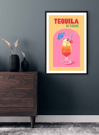 Framed Tequila Sunrise Plakat poster on a dark wall above a wooden dresser with vases and dried plants.