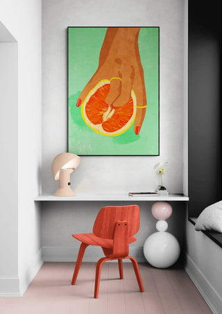 Art print of a hand holding a grapefruit, displayed above a modern desk with a red chair and lamp.