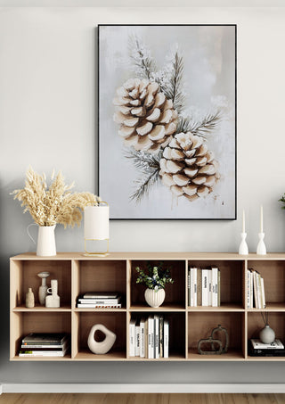 Wall art featuring frosted pine cones above a modern wooden shelf with books, vase, and candle decor.