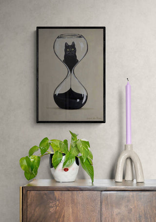 Framed Hourglas Cat - Coco de Paris artwork on wall above cabinet with plant and lilac candle.