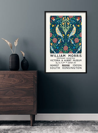 Framed William Morris centenary exhibition poster on a dark wall above a wooden dresser with decorative vases.
