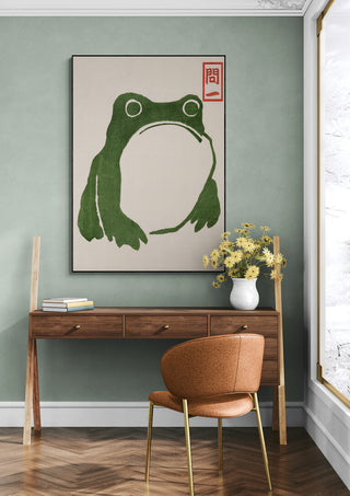 Grumpy Toad II by Matsumoto Hoji framed art above a wooden desk with flowers in a modern room.